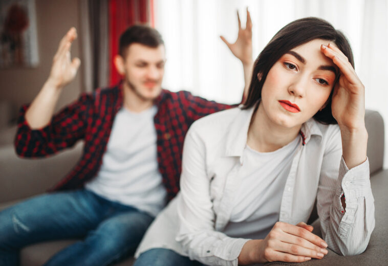 Stressed wife sits on couch, angered husband yells on her, family conflict. Unhappy man and woman in quarrel